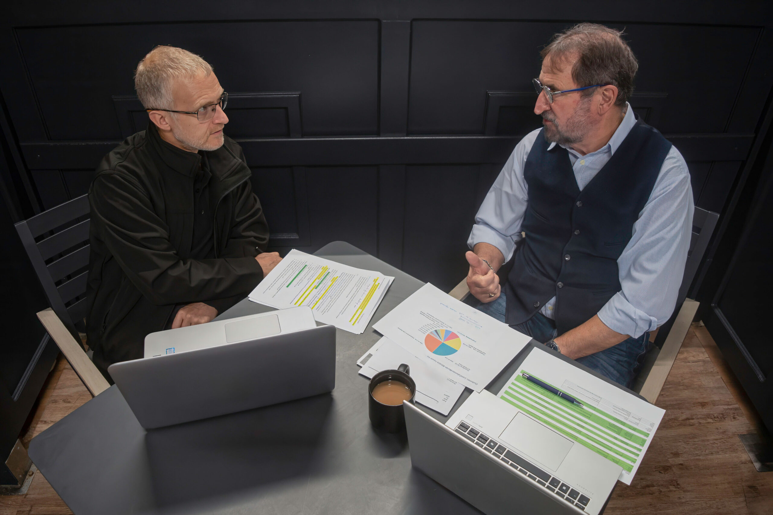 Two men seated at a table, each using a laptop, engaged in conversation and collaboration.