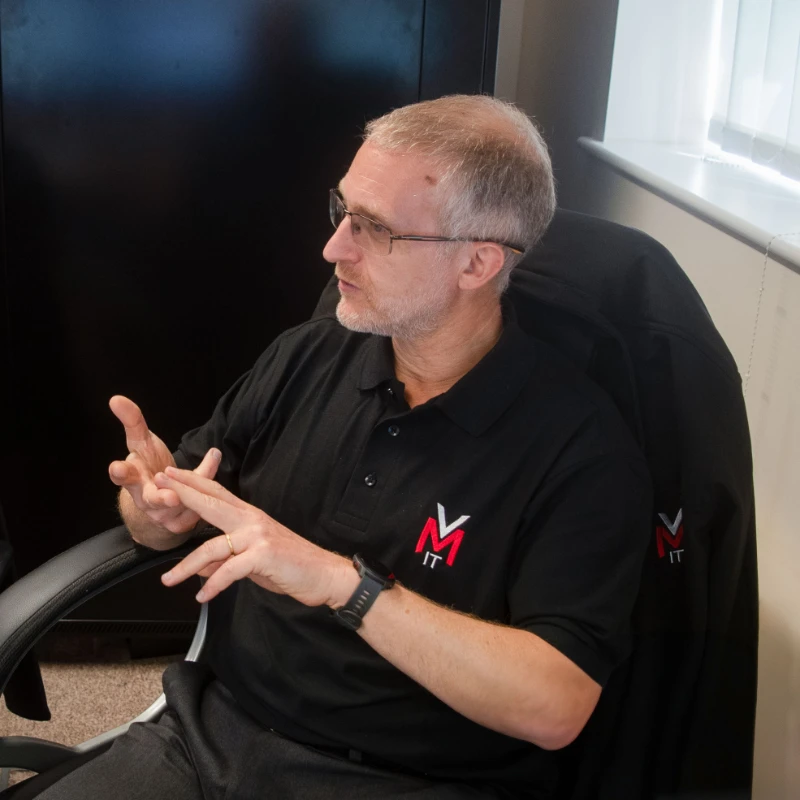 Man wearing a VMIT polo shirt seated in an office chair, speaking and gesturing during a discussion