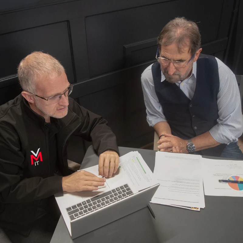Two men reviewing documents and data on a laptop in a business meeting, one wearing an VMit-branded shirt