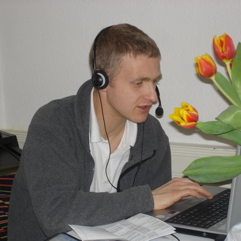 A man wearing headphones is sitting indoors at a desk, using a laptop.