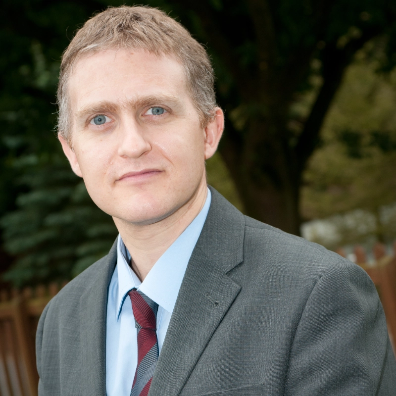 A man in a suit standing outdoors next to a tree, wearing a blazer, dress shirt, and necktie.