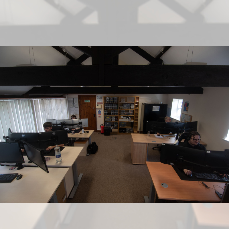A group of people seated at desks working on computers in a modern office space with shelves and an organized interior design.